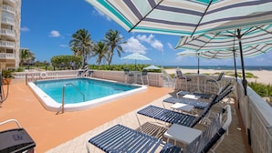 A pool area with lounge chairs and umbrellas, a clear view of the beach, and a high-rise building in the background. - DIRECTLY ON BEACH! JUST STEPS TO THE OCEAN Free Parking Full Kitchen Heated Pool (Pompano Beach)
