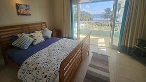 Un dormitorio con una cama de madera, una mesita de noche, un cuadro y vista al océano a través de un ventanal.