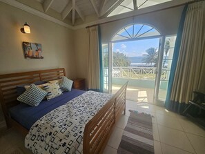 A bedroom with a wooden bed, a nightstand, a painting, and a view of the ocean through a glass door.