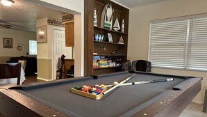 Ein Billardtisch mit Kugeln und Spielstangen, ein Holzregal mit Dekorationsobjekten sowie ein Fenster mit Jalousien.