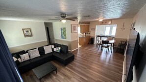 Ein Wohnzimmer mit Ecksofa, einem Esstisch mit Stühlen, einer Küchenzeile und einem Deckenventilator.