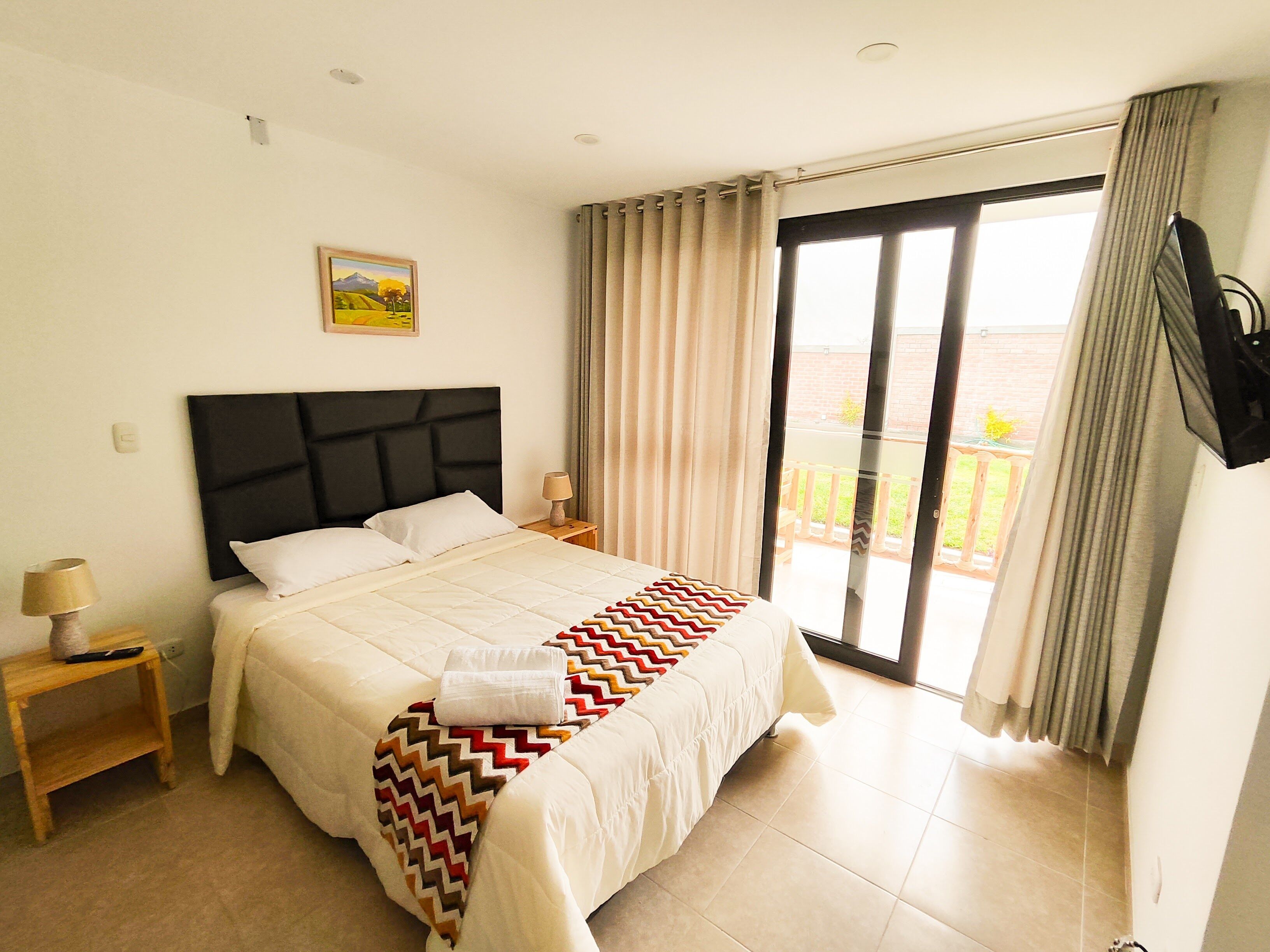 A hotel room with a bed, a nightstand, a lamp, a painting, and a view of the outdoors through a sliding door.
