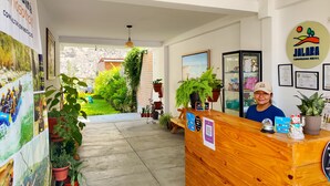 A hotel reception desk with a wooden counter, a person standing behind it, and a view of a garden with potted plants and a brick wall. - Jalara Lunahuaná Hotel (Lunahuana)