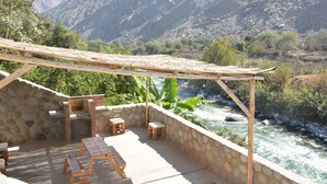 An outdoor patio with a stone wall, wooden furniture, and a view of a river and mountains. - Jalara Lunahuaná Hotel (Lunahuana)