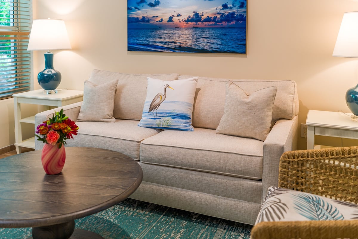 Ein Wohnzimmer mit einem beigefarbenen Sofa, einem runden Couchtisch, einem Gemälde, das einen Sonnenuntergang über dem Meer zeigt, und zwei Lampen.