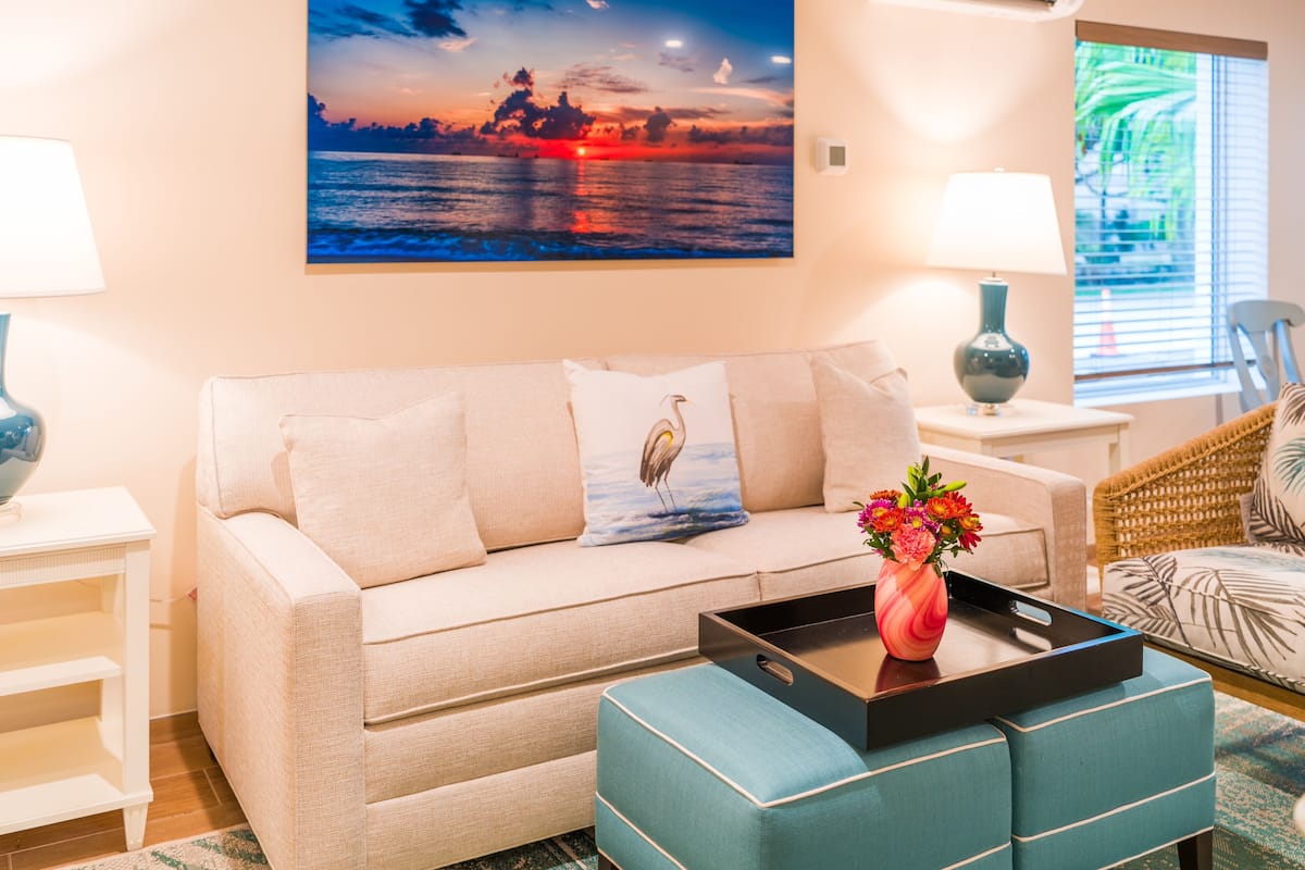 Ein Wohnzimmer mit einem beigen Sofa, einem türkisfarbenen Hocker, einem Sonnenuntergangsgemälde, einer Vase mit Blumen und einer Ablagefläche auf dem Hocker.