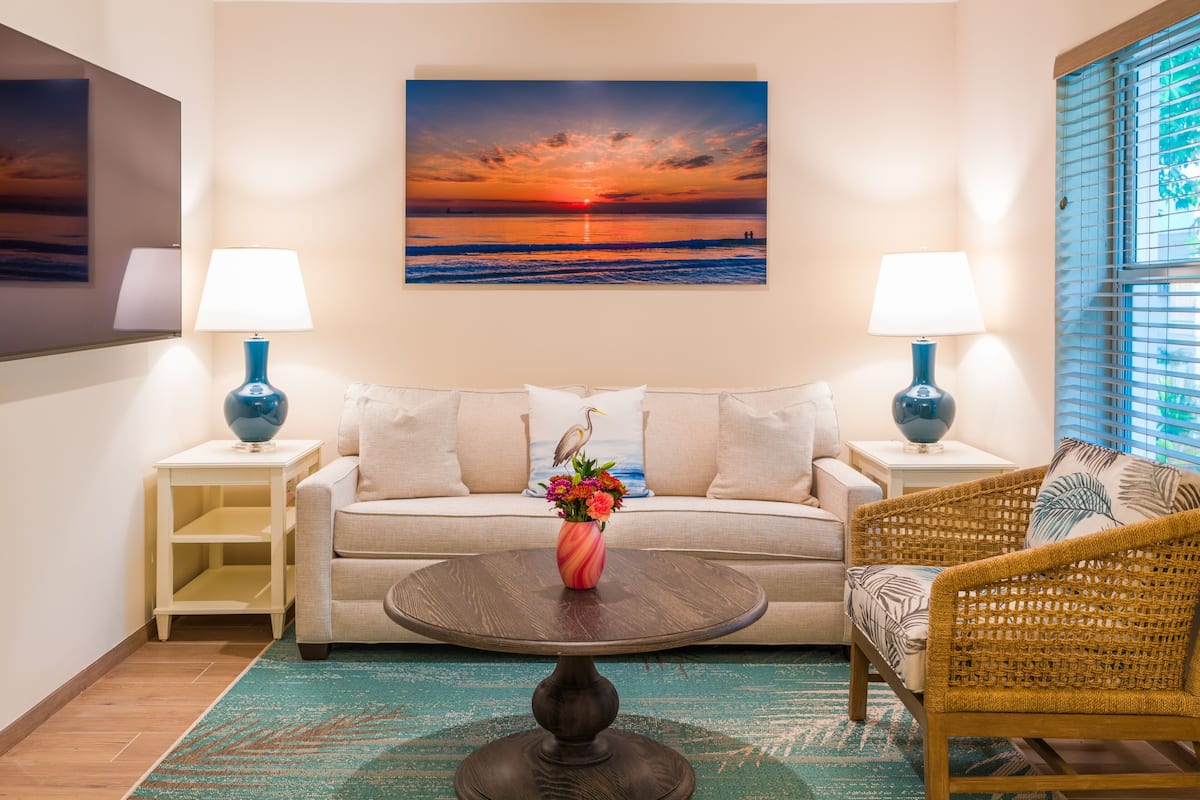 Ein Wohnzimmer mit einer Couch, einem Korbsessel, einem Couchtisch mit Blumenvase und einem Gemälde, das einen Sonnenuntergang über dem Meer zeigt.