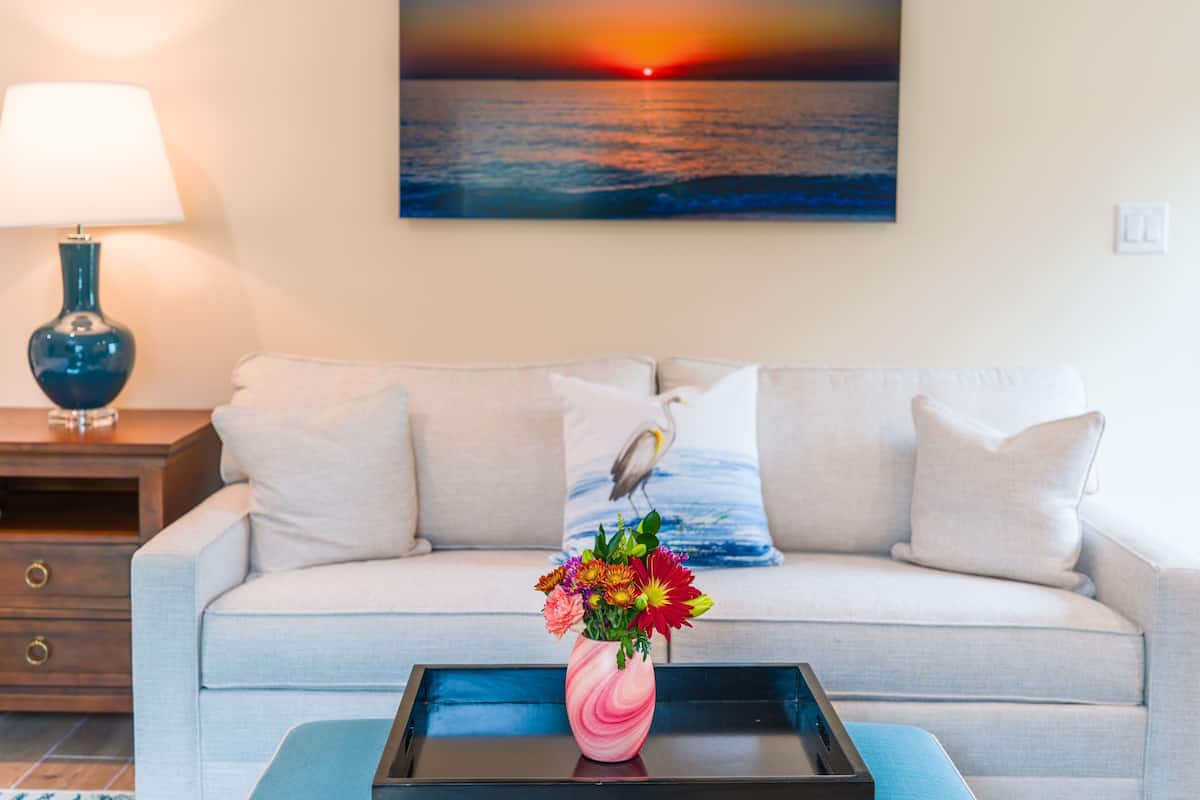 Ein Wohnzimmer mit einer Couch, einem Couchtisch mit Blumenvase, einer Lampe und einem Gemälde, das einen Sonnenuntergang über dem Meer zeigt.