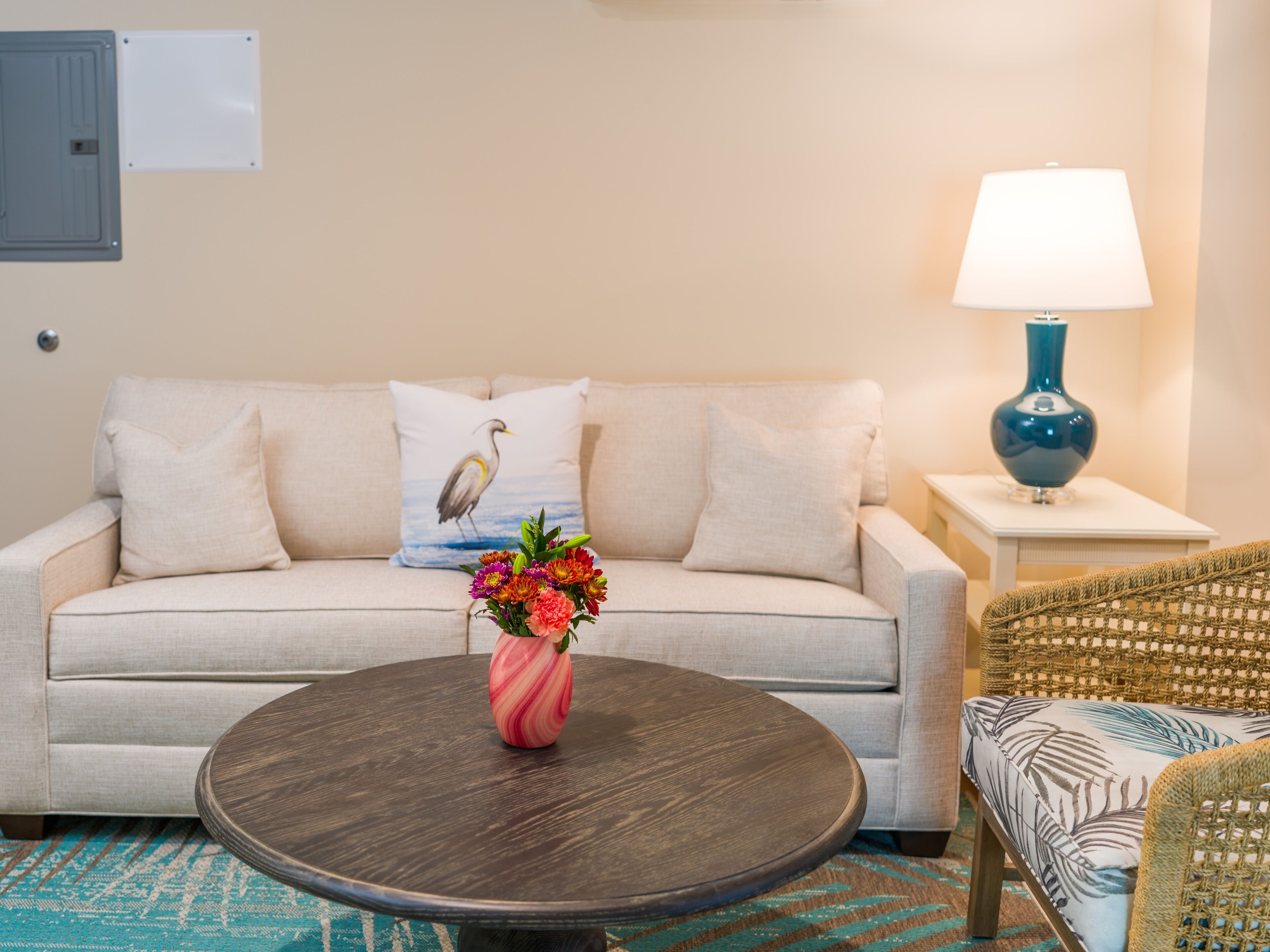 A living room with a beige sofa, a round coffee table with a pink vase, a blue lamp, and a wicker chair.