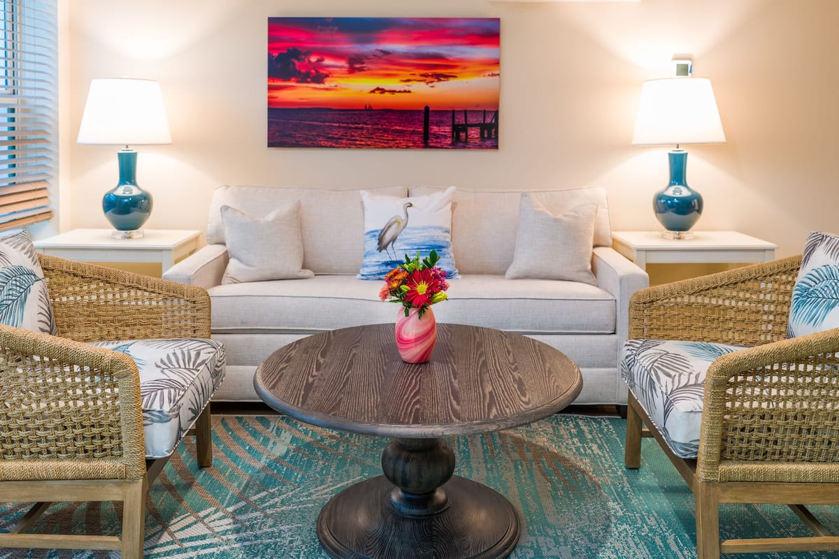 Ein Wohnzimmer mit einer Couch, zwei Beistelltischen mit Lampen, einem runden Couchtisch mit einer Blumenvase und einem Gemälde, das einen Sonnenuntergang über dem Meer zeigt.