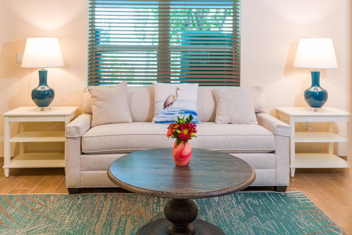Ein Wohnzimmer mit einem beigefarbenen Sofa, zwei Nachttischen mit Lampen, einem runden Couchtisch mit Blumen und einem gemusterten Teppich.