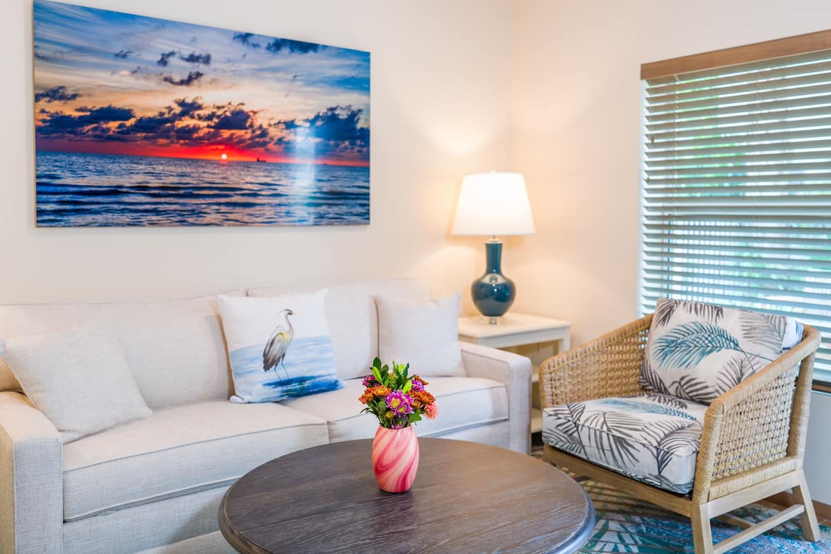 Ein Wohnzimmer mit einer Couch, einem Sessel, einem runden Tisch mit Blumenvase, einer Lampe und einem Gemälde, das einen Sonnenuntergang über dem Meer zeigt.