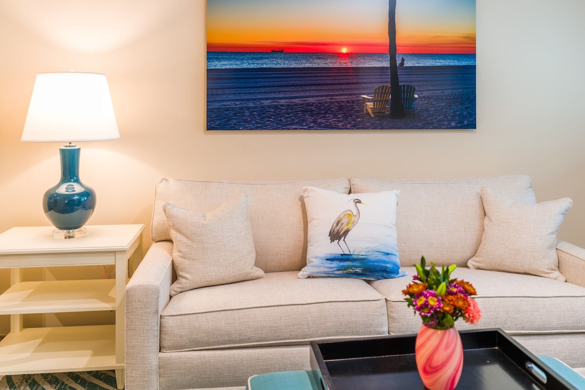 Ein Wohnzimmer mit einem beigen Sofa, einem Gemälde, das einen Sonnenuntergang über dem Meer zeigt, einer Lampe und einer Vase mit Blumen.