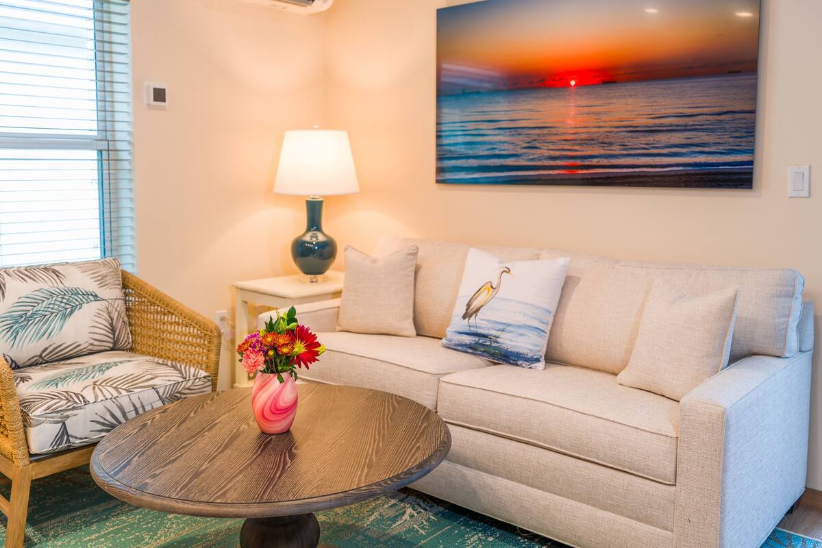 Ein Wohnzimmer mit einer Couch, einem Couchtisch mit Blumen und einer Lampe, sowie einem Gemälde, das einen Sonnenuntergang über dem Meer zeigt.