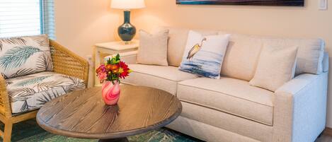 A living room with a sofa, a coffee table with a vase of flowers, a lamp, and a painting of a sunset over the ocean.