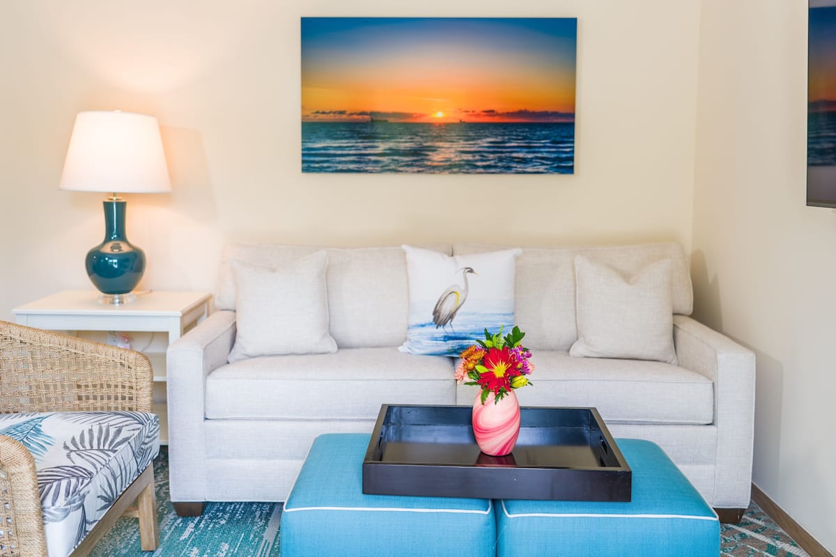 Ein Wohnzimmer mit einer Couch, einem Couchtisch mit Blumenvase, einer Lampe und einem Gemälde, das einen Sonnenuntergang über dem Meer zeigt.