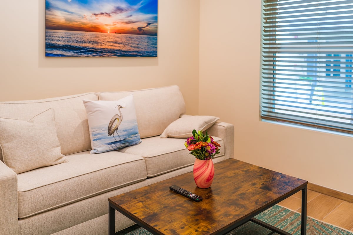 Ein Wohnzimmer mit einem beigefarbenen Sofa, einem Couchtisch mit Blumenvase, einem Gemälde, das einen Sonnenuntergang über dem Meer zeigt, und einem Fenster mit Jalousien.