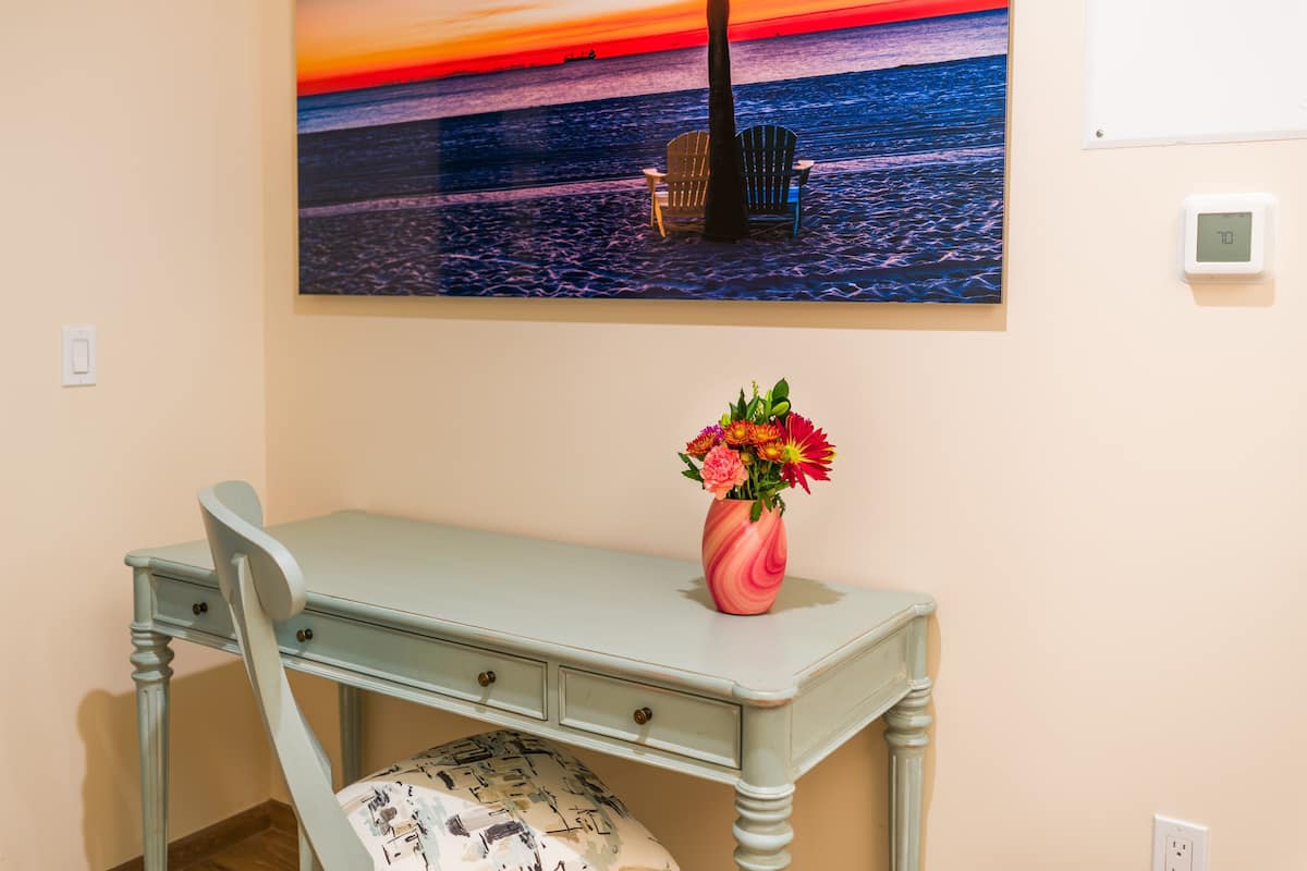 Ein Zimmer mit Schreibtisch, Stuhl, Blumen in einer Vase und einem Gemälde, das einen Sonnenuntergang über dem Meer zeigt.