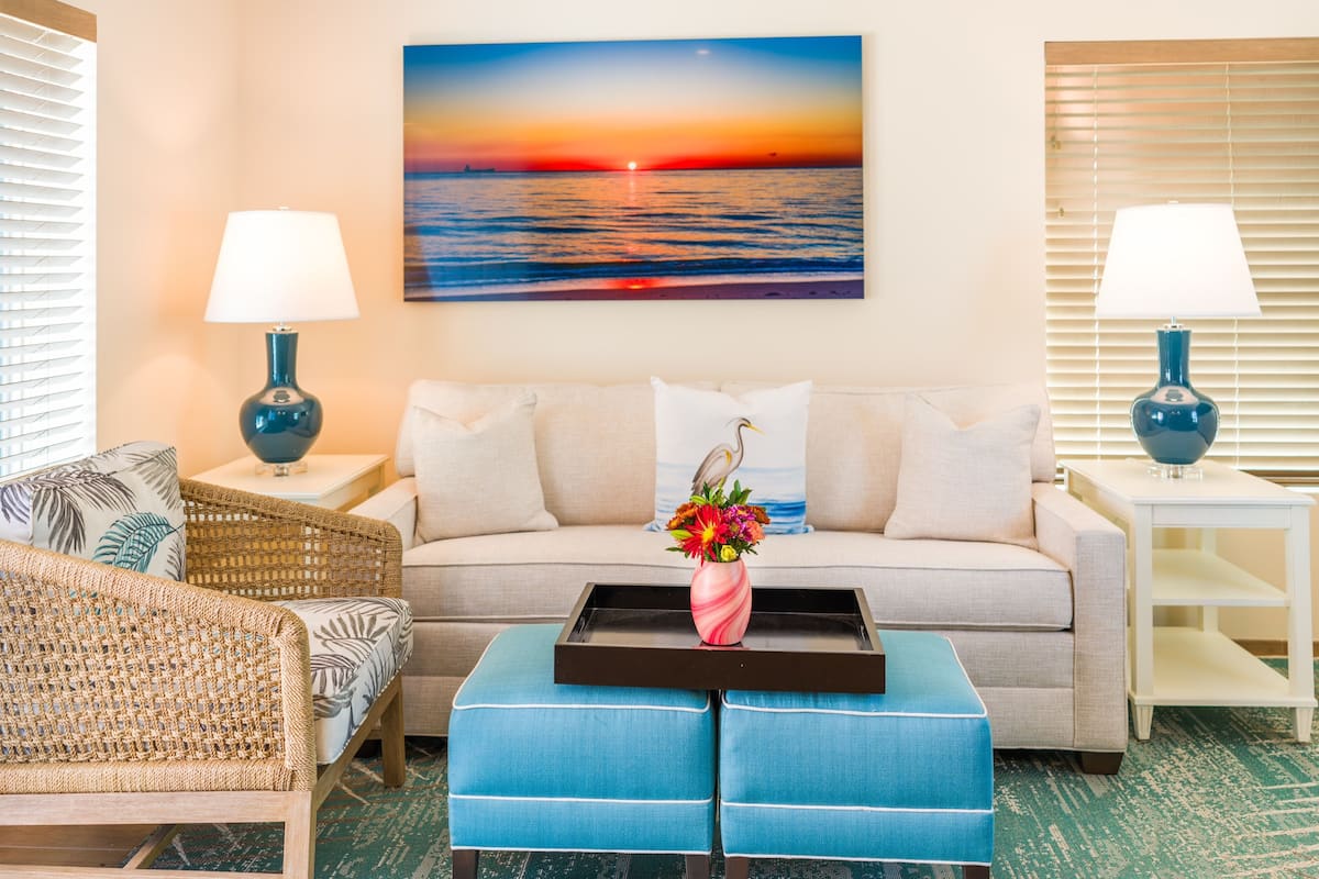 Ein Wohnzimmer mit einer Couch, zwei Nachttischen mit Lampen, einem Korbstuhl, einem blauen Hocker und einem Wandbild, das einen Sonnenuntergang über dem Meer zeigt.