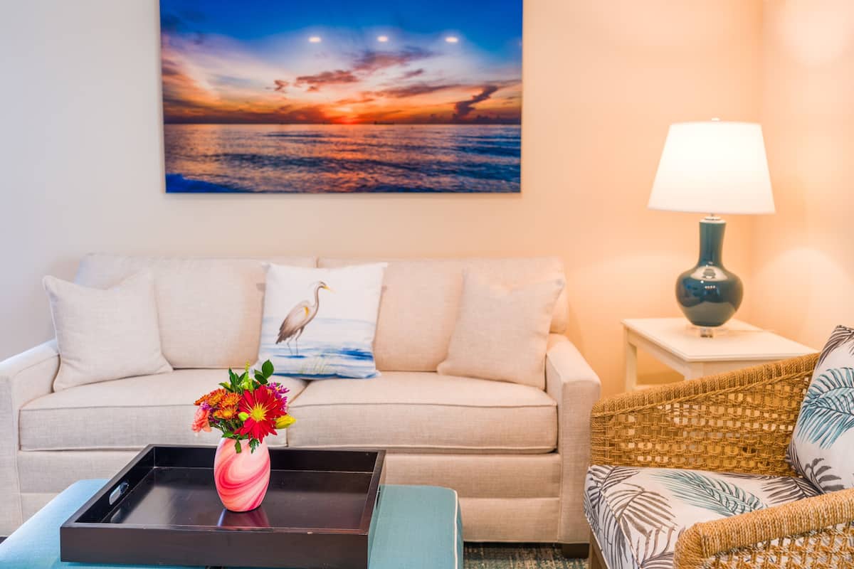 Ein Wohnzimmer mit einer Couch, einem Couchtisch mit Blumen, einer Lampe und einem Gemälde, das einen Sonnenuntergang über dem Meer zeigt.
