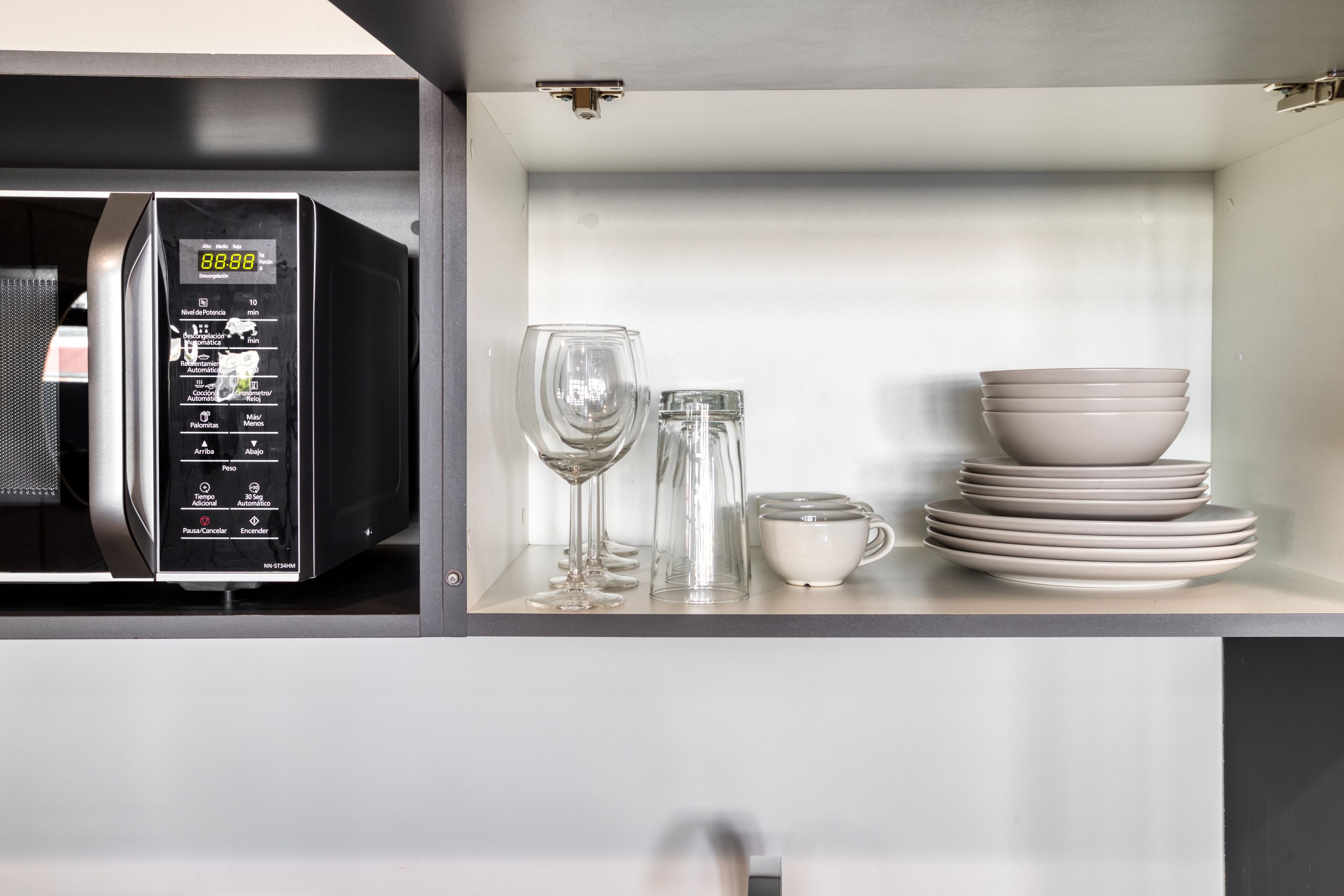A microwave with a digital display, a glass, a glass carafe, and stacked plates and bowls on a shelf.