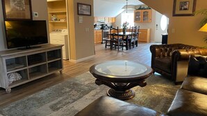 A living room with a television, a glass-top coffee table, a leather sofa, and a dining area with wooden cabinets and a chandelier.