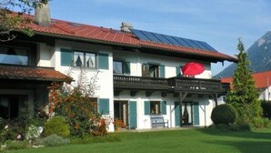 A two-story house with a red roof, a balcony with a red umbrella, and a well-maintained garden.