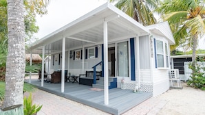 A small, single-story house with a covered porch, a staircase leading to the entrance, and a small garden area with palm trees.