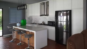 Een moderne keuken met witte kasten, een zwarte koelkast, een gasfornuis en een groene waterdispenser. Er staan drie barkrukken en is een groot raam.
