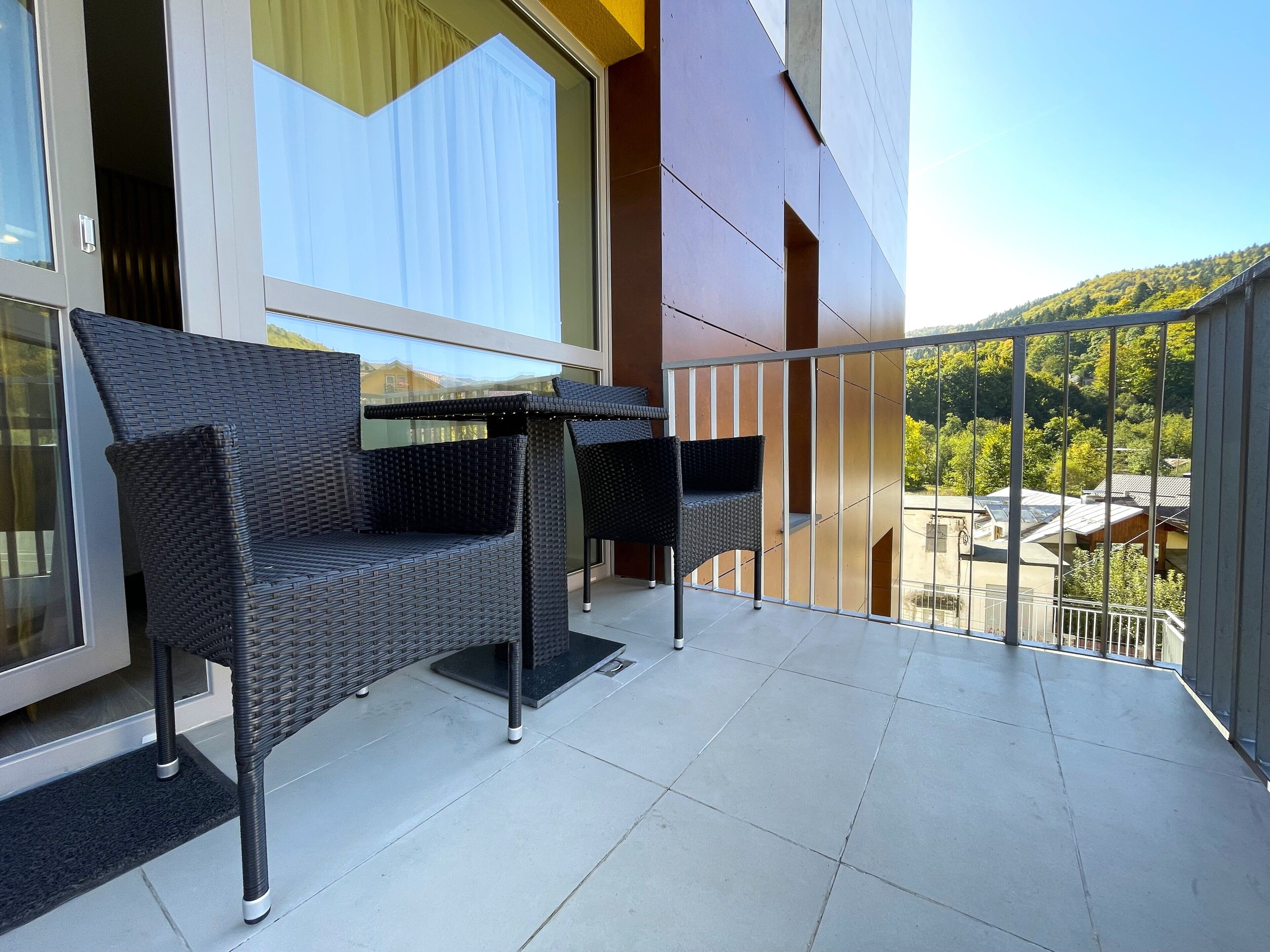 Ein Balkon mit Korbmöbeln, einer Glastür und Blick auf einen grünen Hügel.