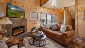 A cozy log cabin interior with a stone fireplace, a large flat-screen TV, a brown leather sofa, and a round coffee table. - SummerFun! Pets, Indoor Pool, Fire Pit, Hot Tub, Foosball/Air Hockey, Dollywood! (Sevierville)