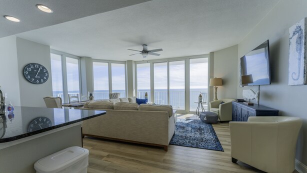Un vaste espace de vie avec une grande fenêtre donnant sur la mer, un ventilateur de plafond, un téléviseur à écran plat et un coin salon confortable.
