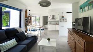 Un salon moderne avec un canapé, une table basse et un téléviseur posé sur un meuble en bois. On y trouve également un coin repas avec une table et des chaises, ainsi qu’une cuisine équipée d’une cuisinière et de placards.