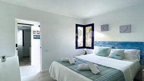 Une chambre à coucher comprenant un lit, une table de chevet, une fenêtre donnant sur des palmiers et une porte menant à la salle de bain.