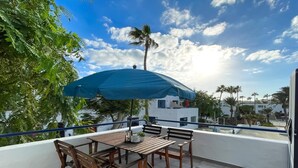 Une terrasse avec une table et des chaises, abritée par un parasol bleu, d’où l’on peut admirer un quartier résidentiel bordé de palmiers et de maisons.