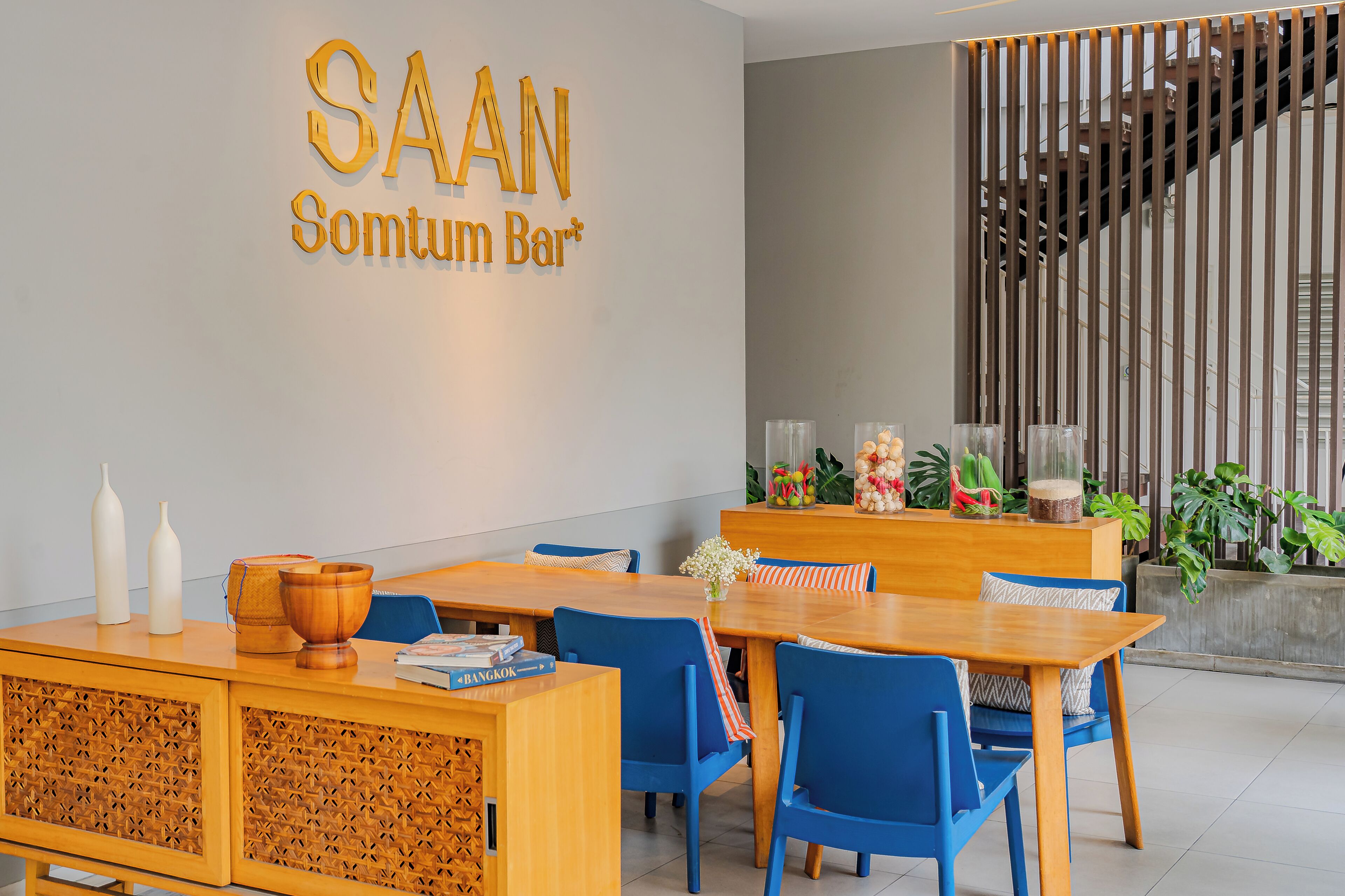 A modern interior with a wooden reception desk, blue chairs, and a wall-mounted sign reading 'SAAN Somtum Bar'.