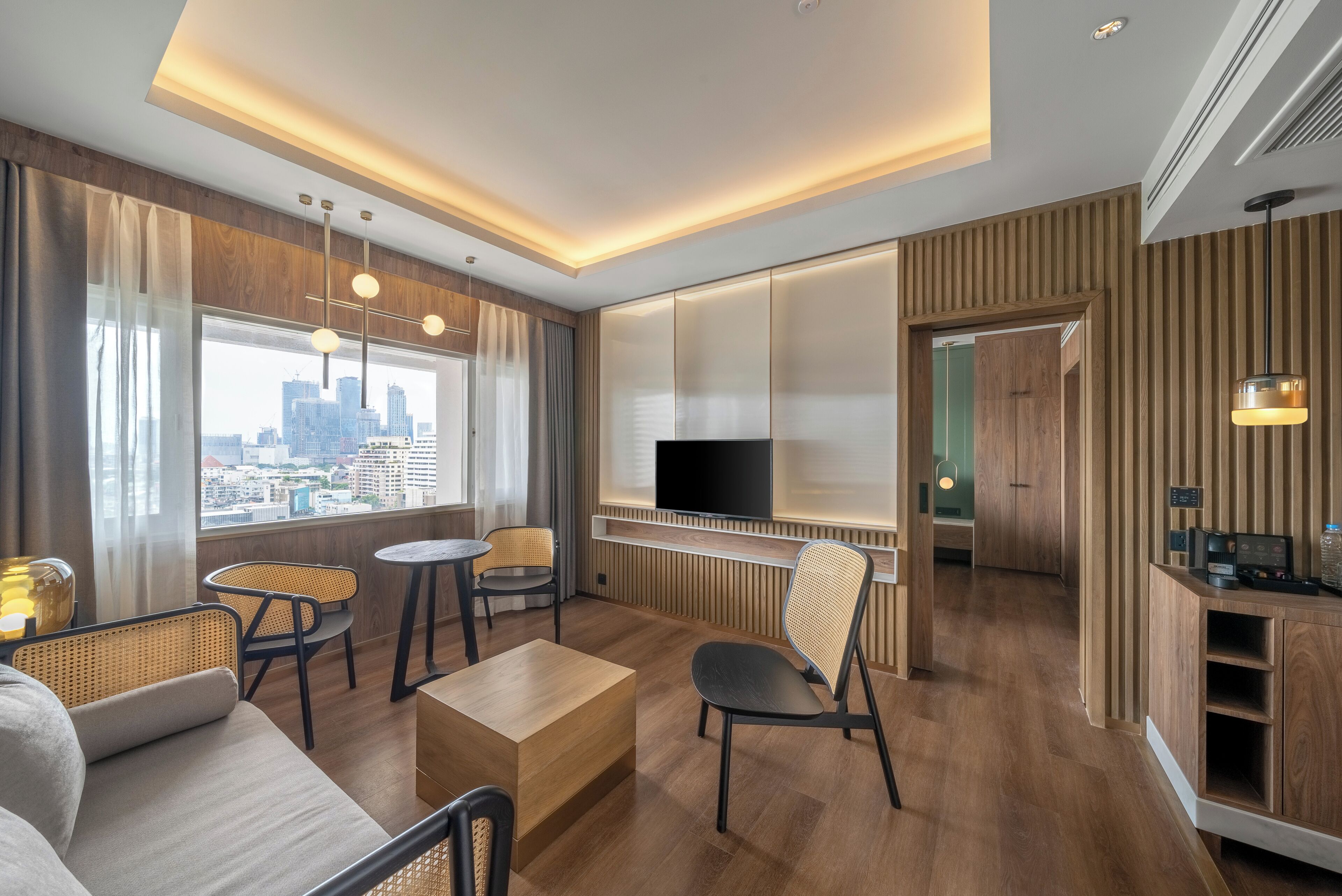 A modern hotel room with a city view, wooden flooring, a flat-screen TV, a dining area, and a kitchenette.