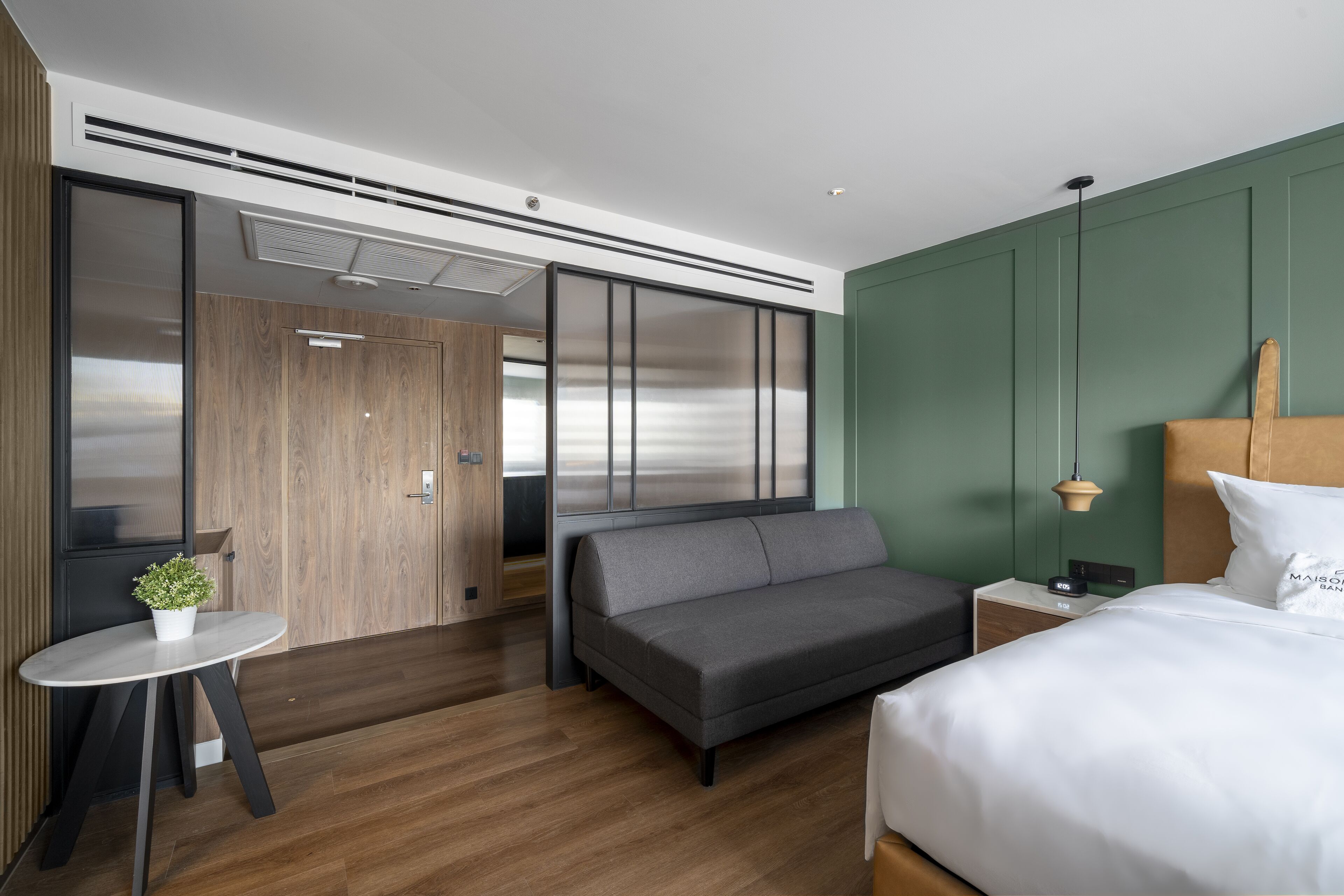 A modern hotel room with a bed, sofa, bedside table, and a small table with a plant.