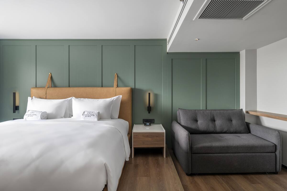 Une chambre d’hôtel moderne avec un grand lit, un fauteuil gris, une table de chevet avec une lampe et un luminaire fixé au mur.