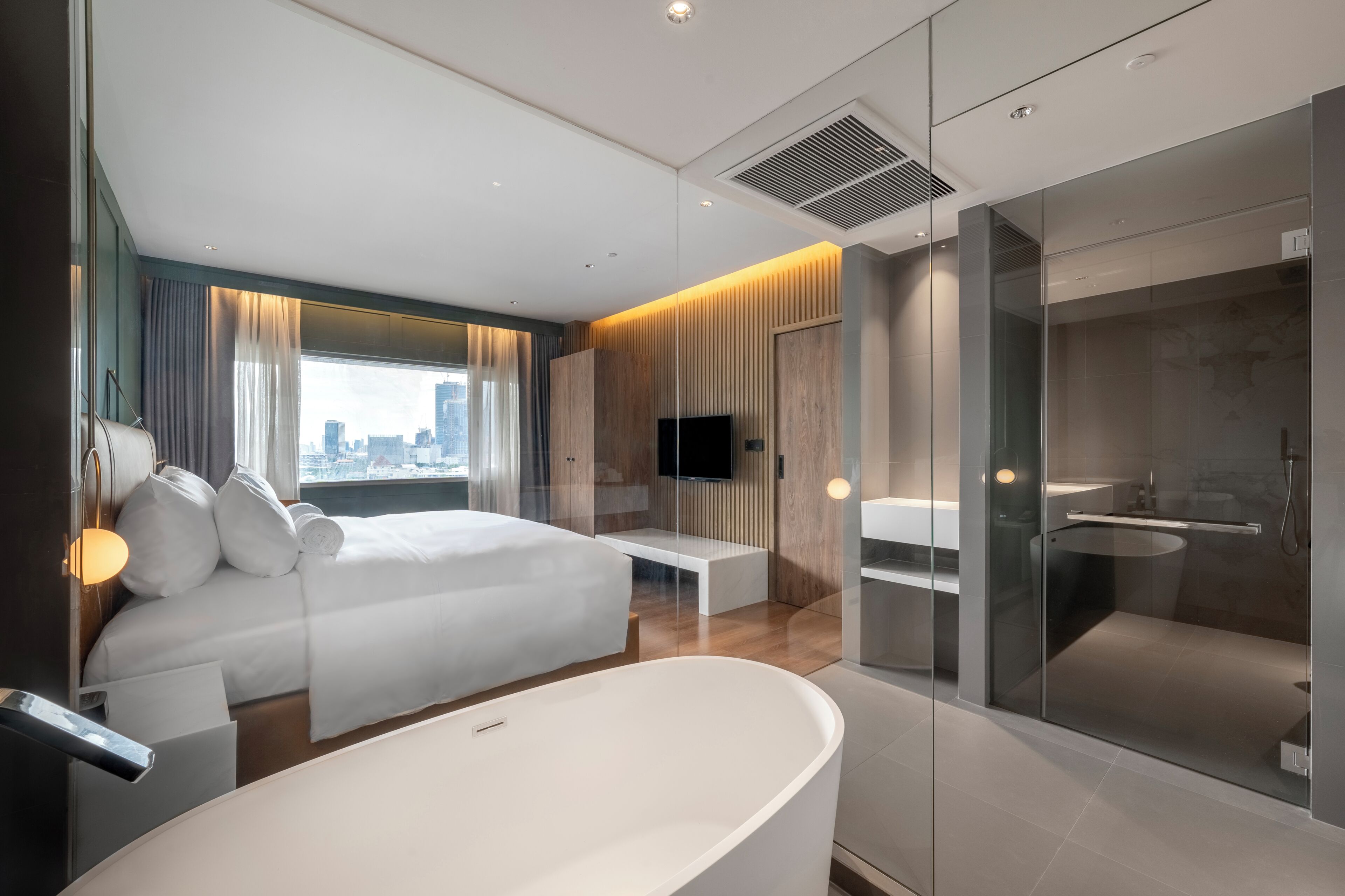 Habitación de hotel moderna con una cama grande, televisión, un banco y zona de ducha con paredes de vidrio.