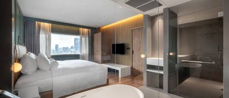 A modern hotel room with a large bed, a TV, a bench, and a glass-enclosed shower area.