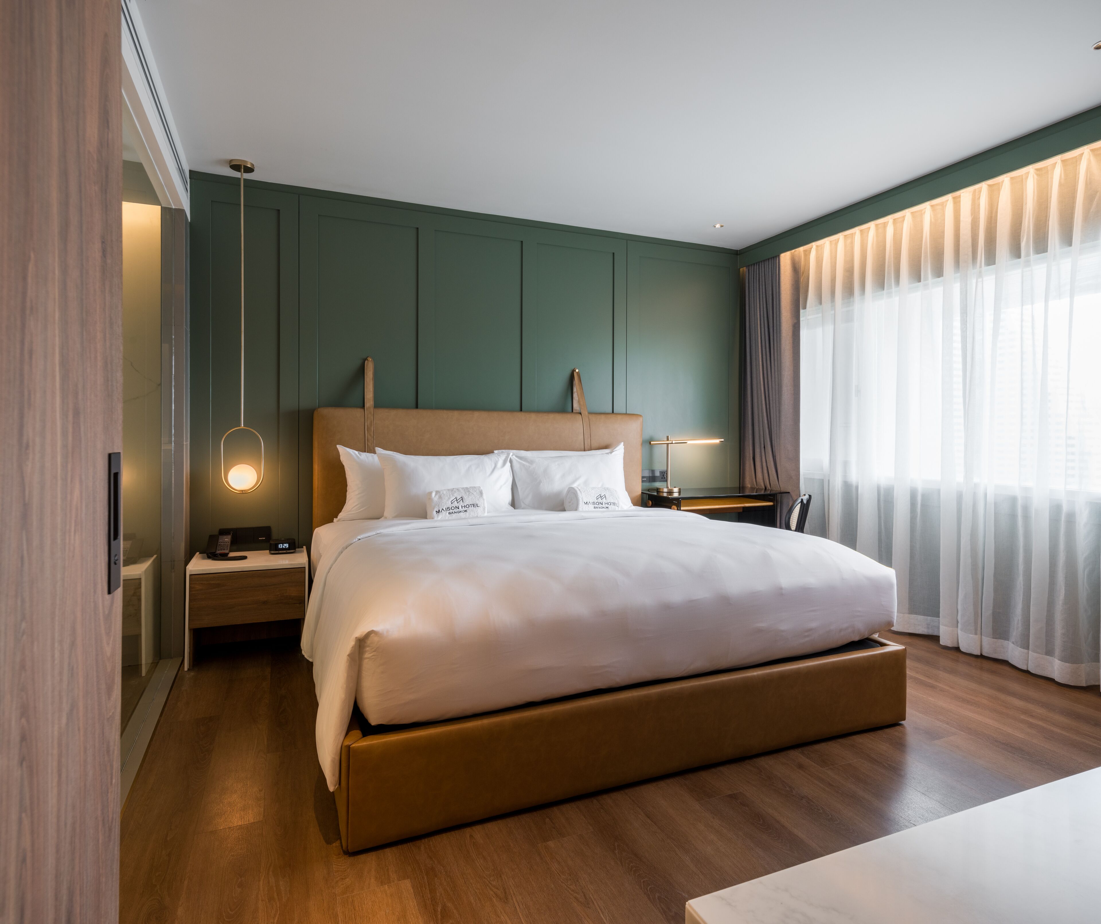 A modern hotel room with a large bed, wooden headboard, bedside tables, and a view of the city through sheer curtains.
