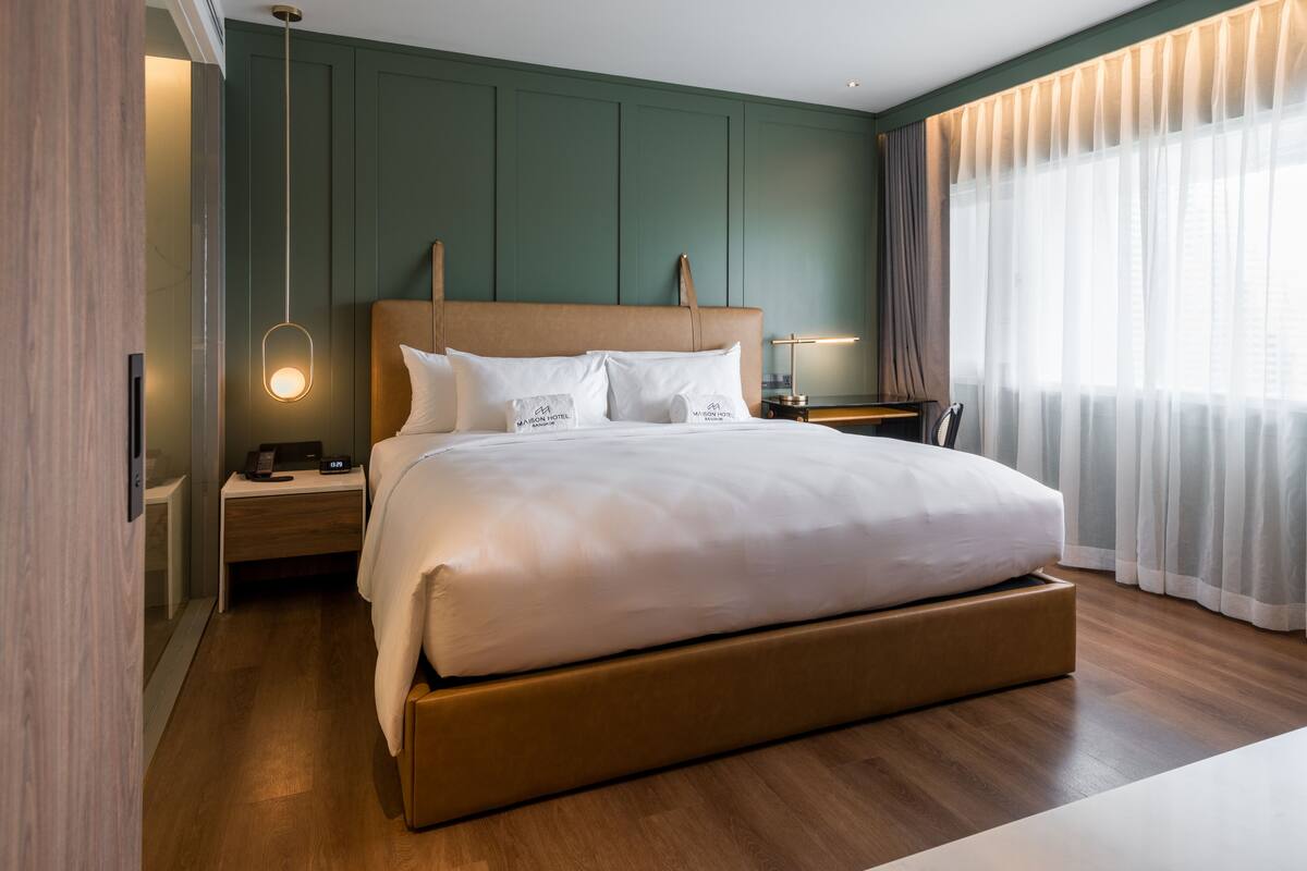 Une chambre d’hôtel moderne dotée d’un grand lit, d’une tête de lit en bois, de tables de chevet et offrant une vue sur la ville à travers de fines rideaux.