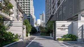 Une entrée d’hôtel moderne, avec un chemin d’accès dégagé menant au bâtiment, entouré de verdure et d’autres immeubles de grande hauteur.