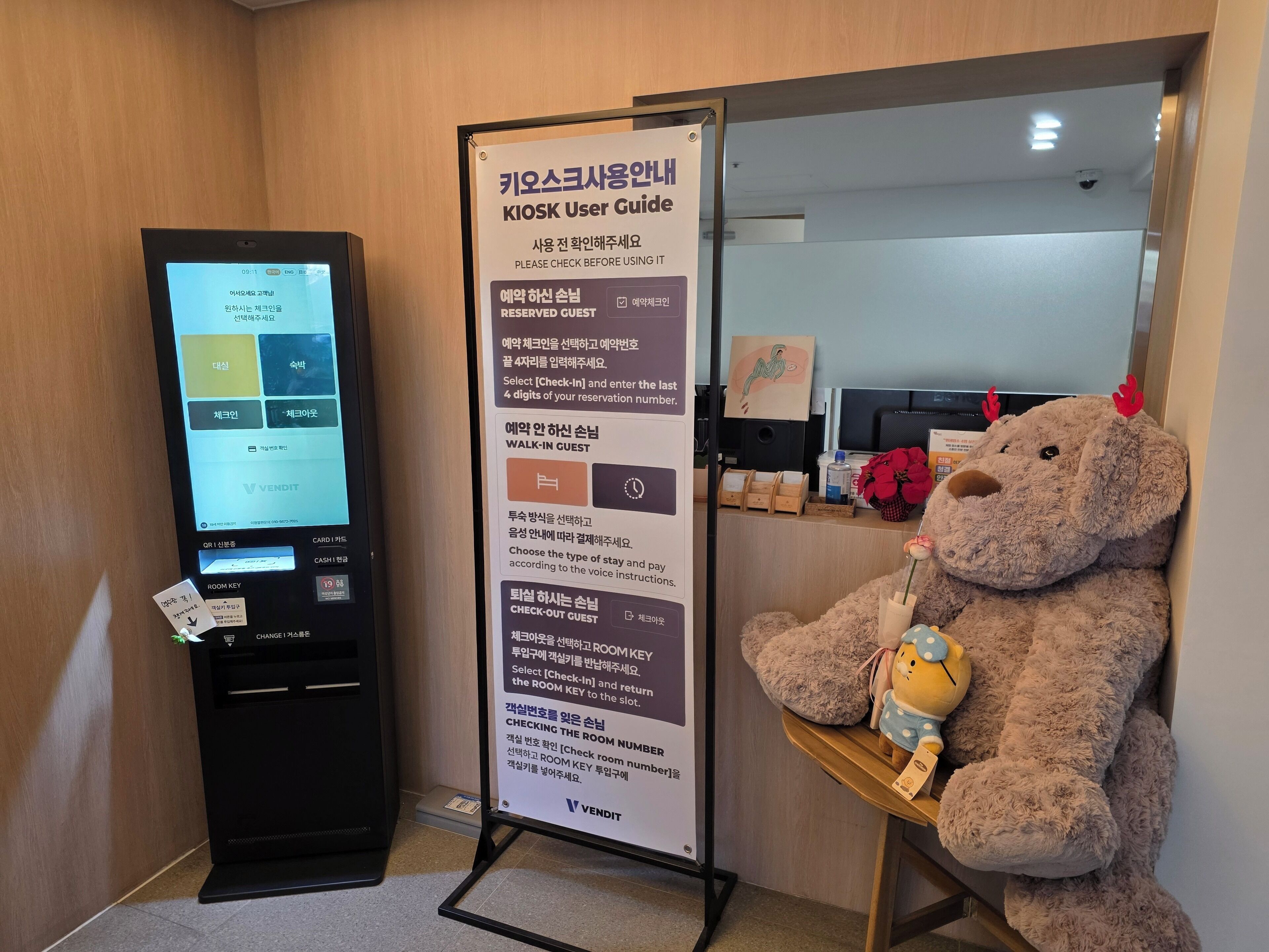 Quiosco con una pantalla mostrando un sistema de reservas, un cartel con instrucciones para usuarios y un oso de peluche en una mesa.