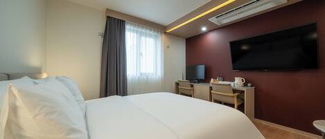 A hotel room with a large bed, a desk with a chair, a flat-screen TV mounted on the wall, and an air conditioning unit.