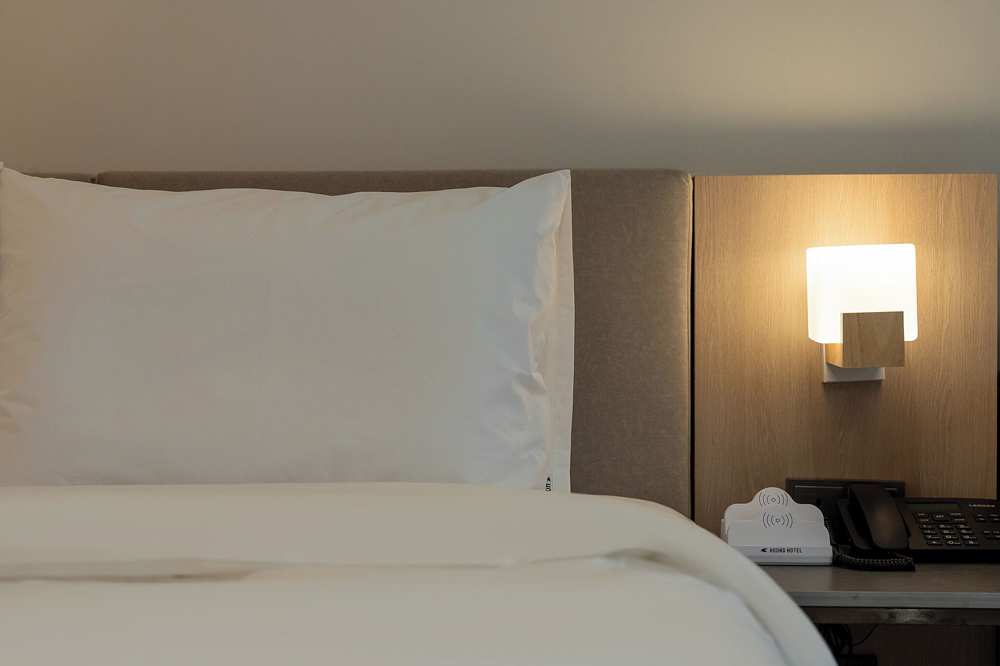 A hotel room with a bed, a bedside table with a lamp, a telephone, and a hotel-branded device.