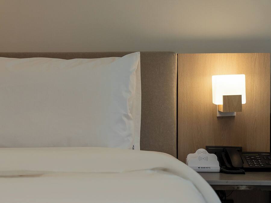 A hotel room with a bed, a bedside table with a lamp, a telephone, and a hotel-branded device.