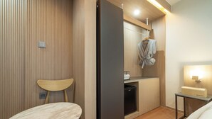 Habitación de hotel pequeña con pared de paneles de madera, una mesa de comedor, un refrigerador negro, una cocina compacta y una mesita de noche con lámpara.