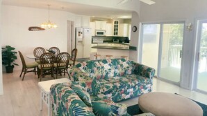 Ein geräumiges Wohnzimmer mit einer großen floralen Couch, einem Essbereich mit Holzsesseln, einer Küche mit weißen Schränken und einer Glastür zum Balkon.
